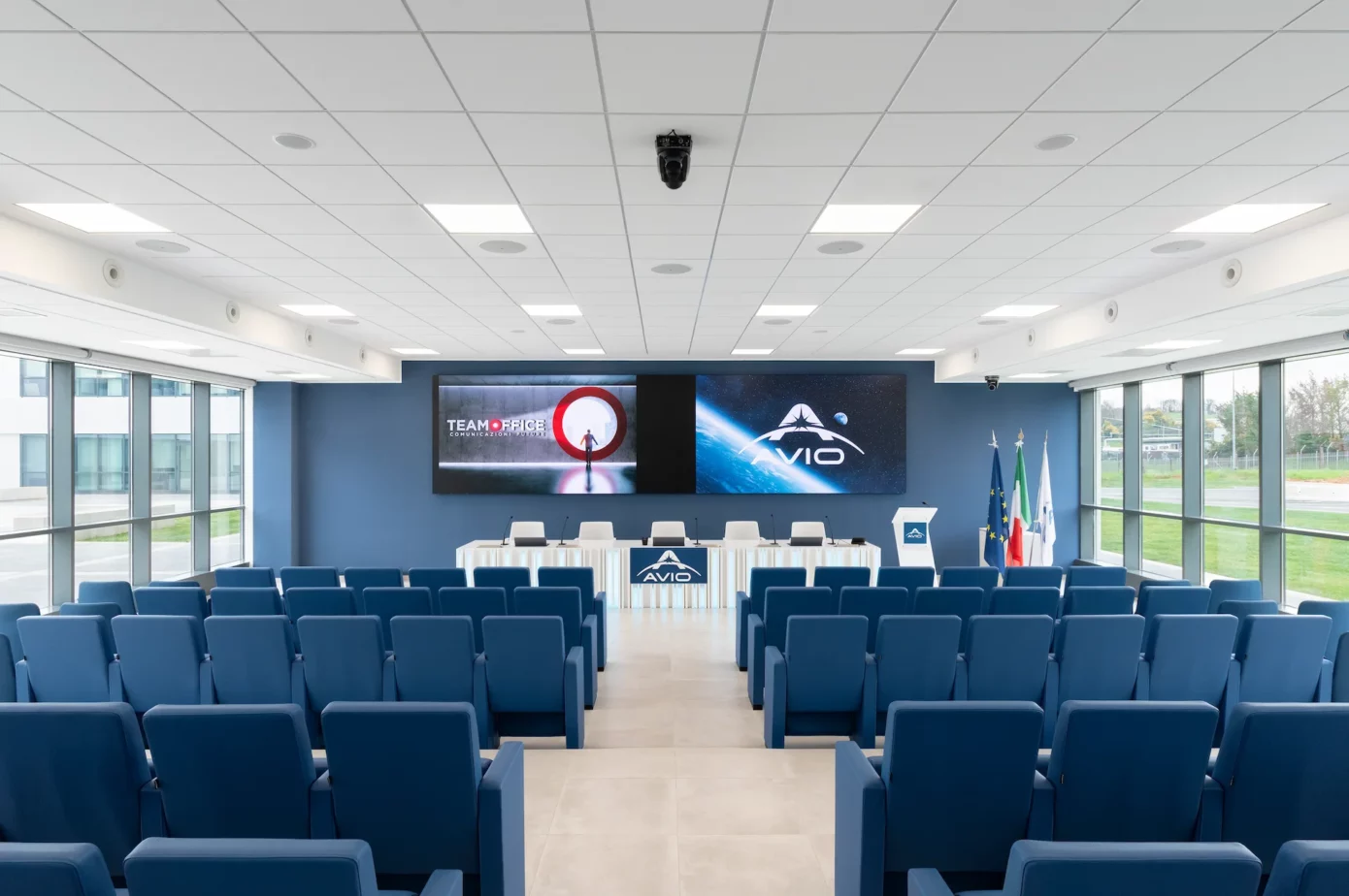 Inquadratura frontale dal retro dell'auditorium. Mostra due file di poltrone blu viste da dietro, mentre in fondo alla sala si vede il tavolo relatori e due ledwall affiancati. La stanza ha lateralmente le pareti finestrate ed è molto luminosa