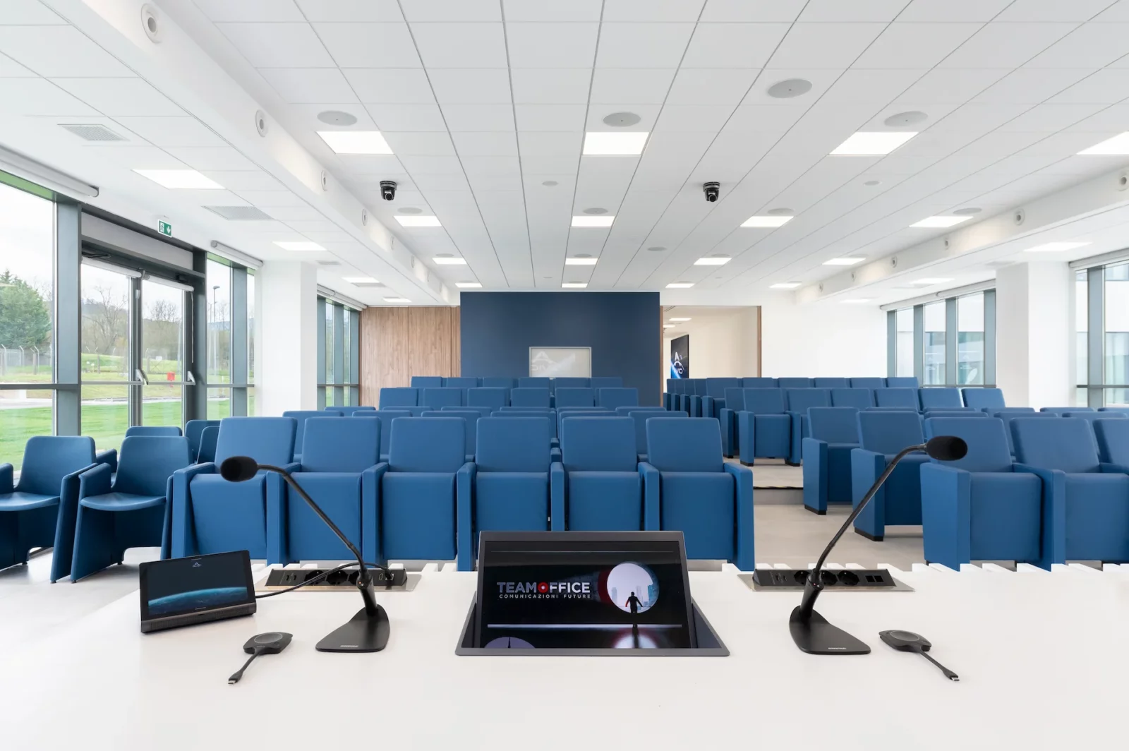 auditorium visto dal banco relatori. in primo piano c'è un monitor incassato a tavolo e i microfoni per i relatori. Dietro il tavolo le varie file di sedute della platea, tutte blu