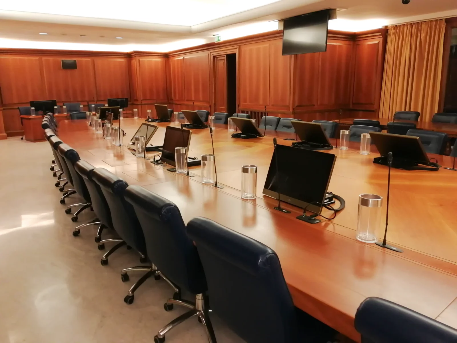 Sala del consiglio con sedute nere eleganti. L'ambiente è caldo e accogliente, con le pareti in legno. In primo piano c'è il tavolo della boardroom in legno. Il tavolo accoglie molte postazioni. Ogni due postazioni è presente un monitor automatizzato, mentre per ogni seduta è disponibile un microfono a collo d'oca installato direttamente a tavolo