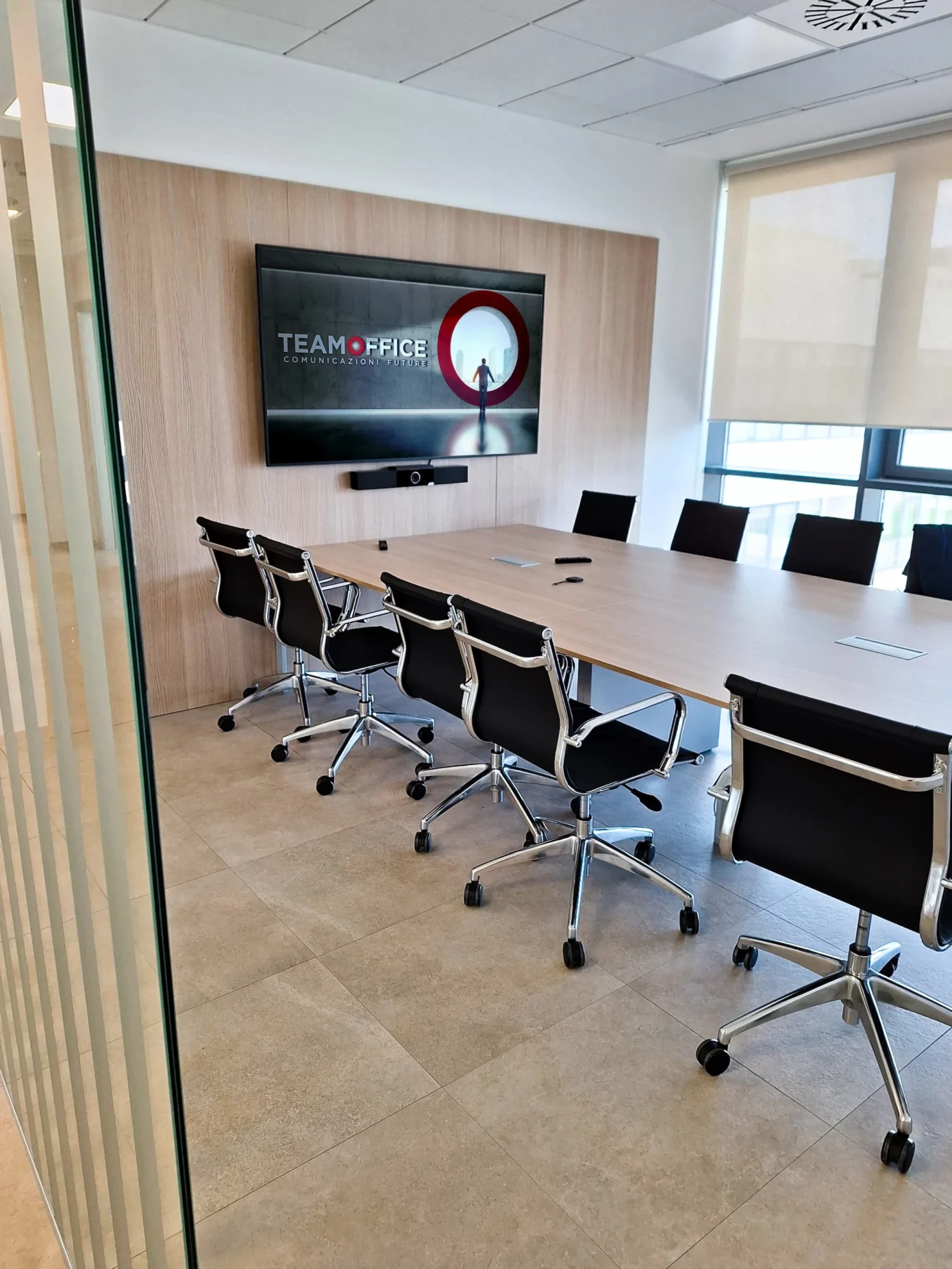 Sala meeting vista lateralmente. Al centro c'è un grande tavolo in legno circondato da sedie nere e alluminio, sulla parete di fondo è installato un monitor ldf con sotto una barra per videoconferenza poly studio