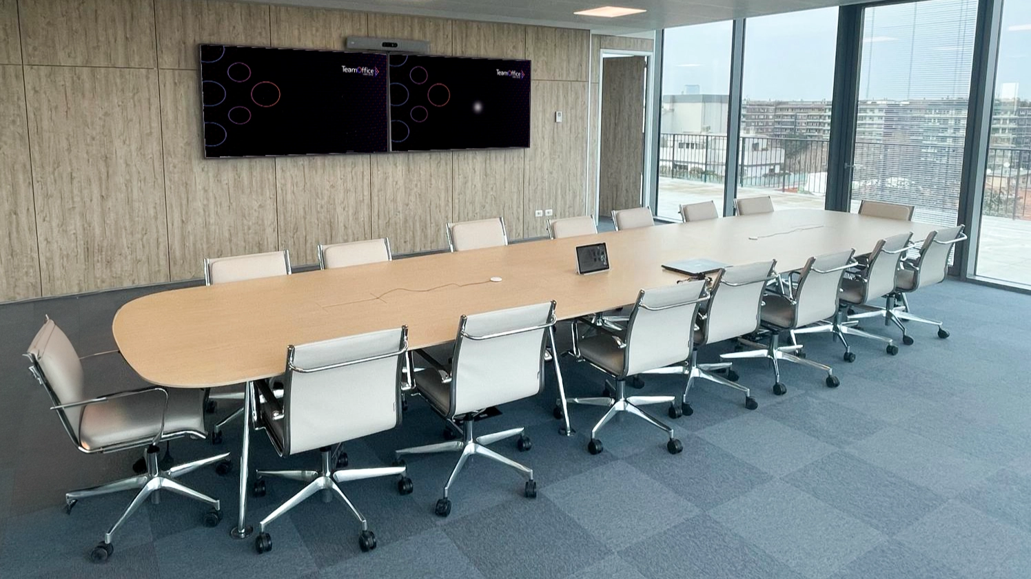 panoramica di una sala riunione grande, con un tavolo in legno su cui è poggiato un pannellino di controllo. Sul muro dietro la sala è installato un doppio monitor con sopra una videobar